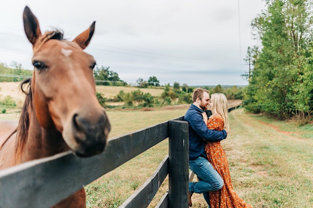 Couple kissing by a horse pasture with a horse looking at the camera