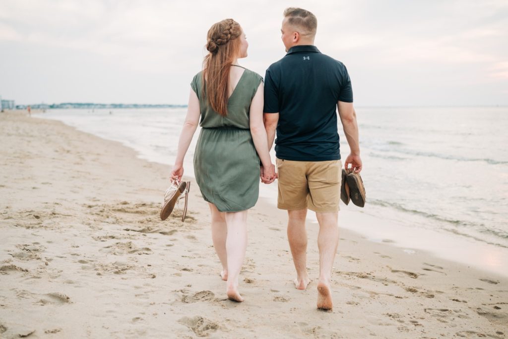 Couple from behind walking down the beach holding hands and carrying their shoes for their Virginia Beach engagement photos