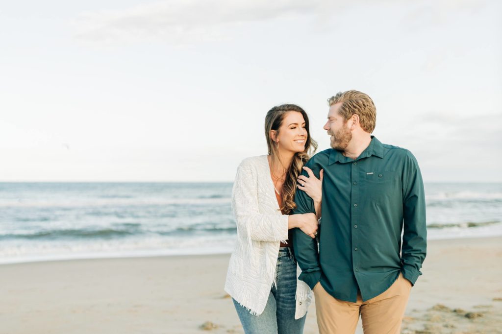 Girl in jeans and orange shirt on the oceanfront walking with her fiancé during sunset taking engagement portraits