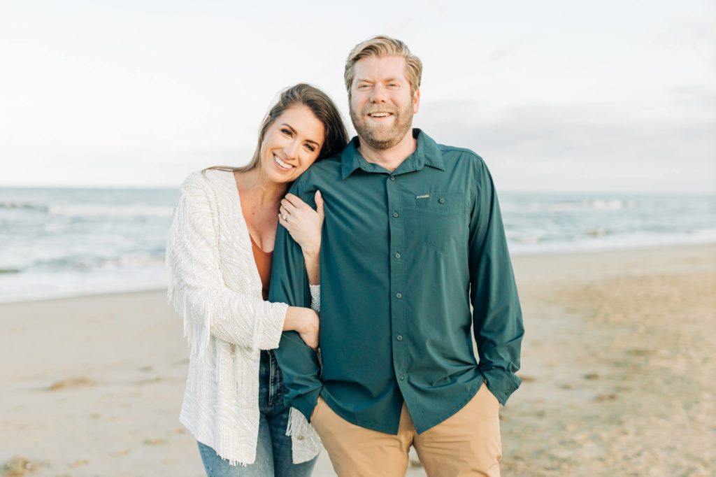 Engaged couple in jeans on the beach smiling at the camera