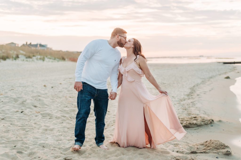 Engaged couple kissing on the beach at sunset on a summer day. Girl wearing pink long dress for engagement photos. 